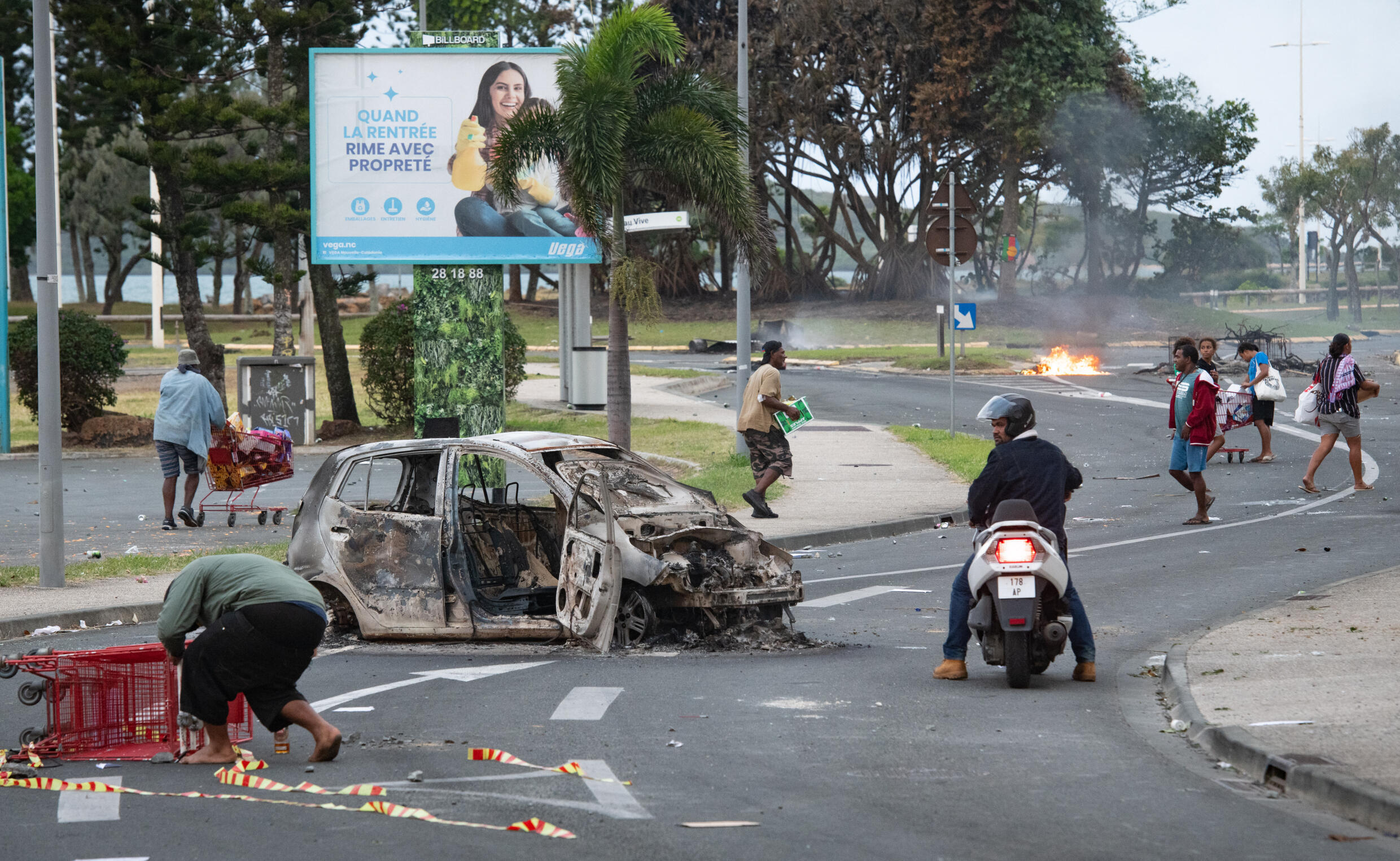 Des gens marchent dans la rue en poussant des caddies à côté d'une voiture incendiée après le pillage d'un supermarché et la vandalisation de magasins dans le quartier N'Gea de Nouméa, le 14 mai 2024.