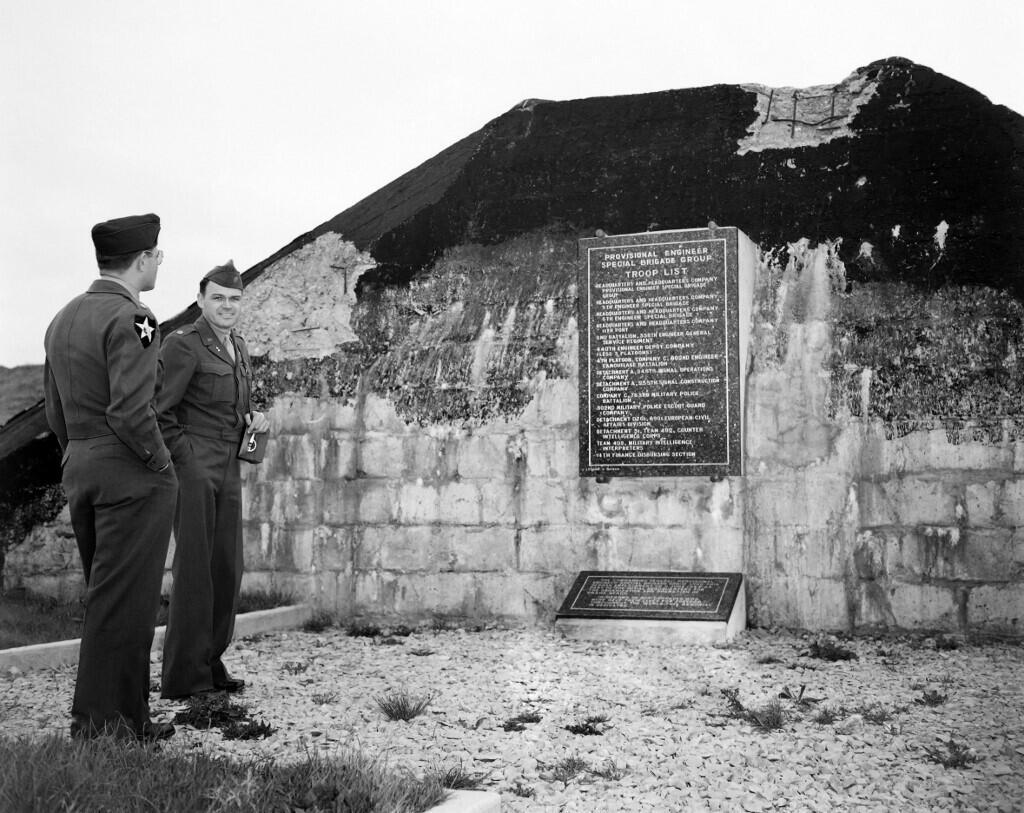 Des hommes en uniforme militaire se tiennent devant un mémorial sur une plage de Normandie en juin 1954, lors des commémorations du 10e anniversaire du Débarquement.